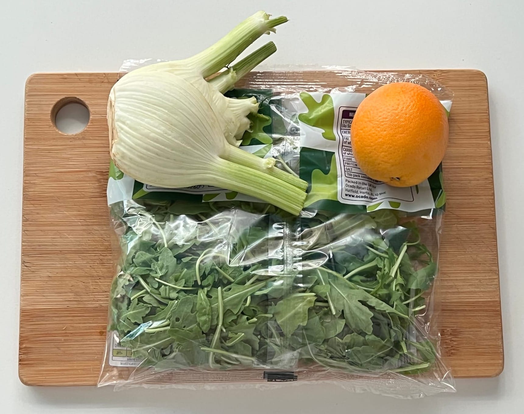 Whole fennel bulb, packaged rocket, and an orange on a wooden chopping board ready for preparation