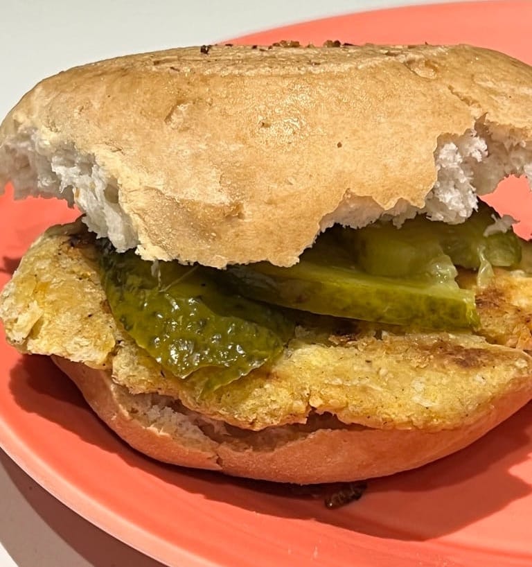 Low-protein burger with pickles in a bun on a coral plate, showing a bite taken from the top bun.