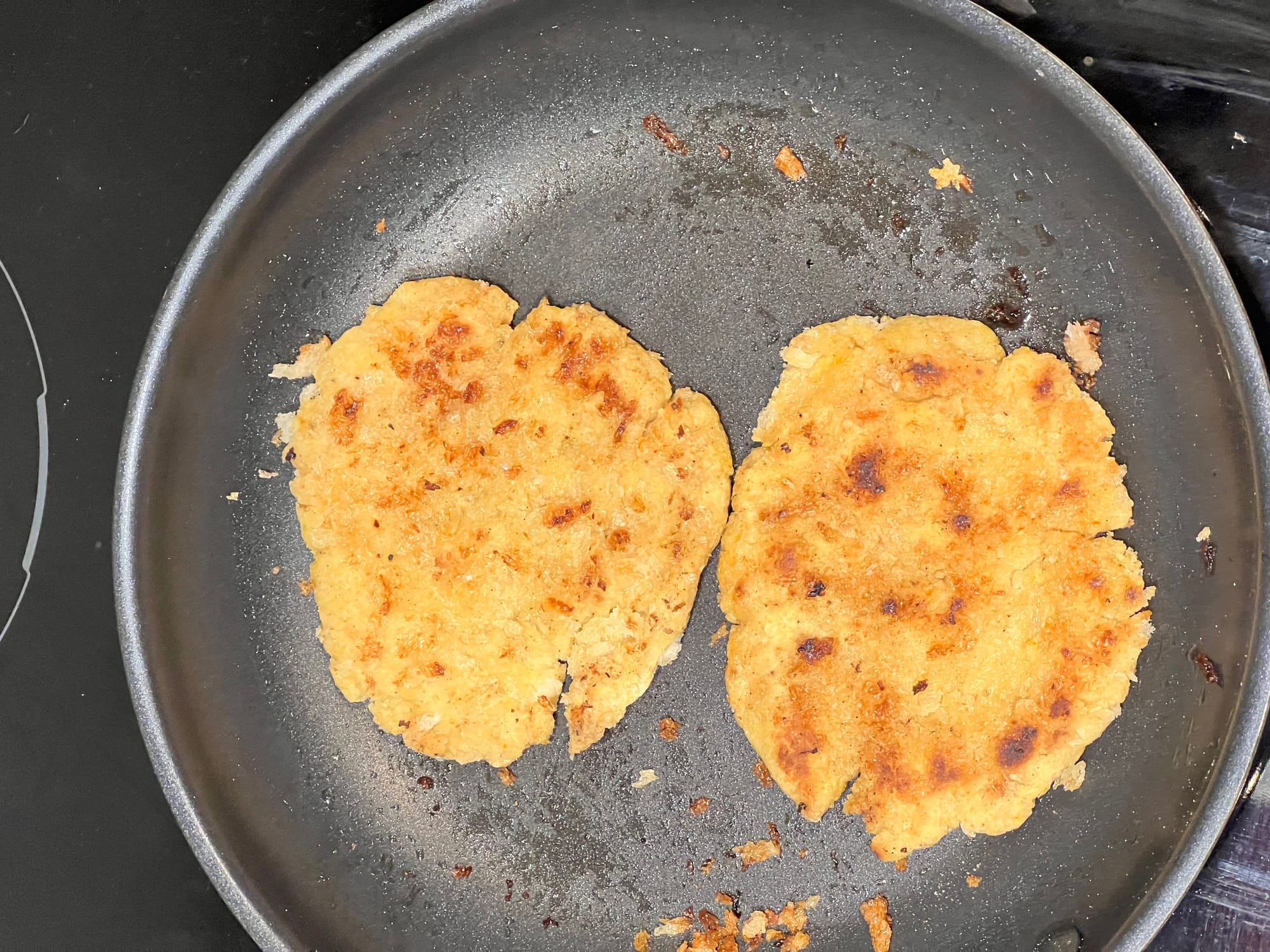 Two golden-brown low-protein patties cooking in a non-stick frying pan, lightly browned on the surface.