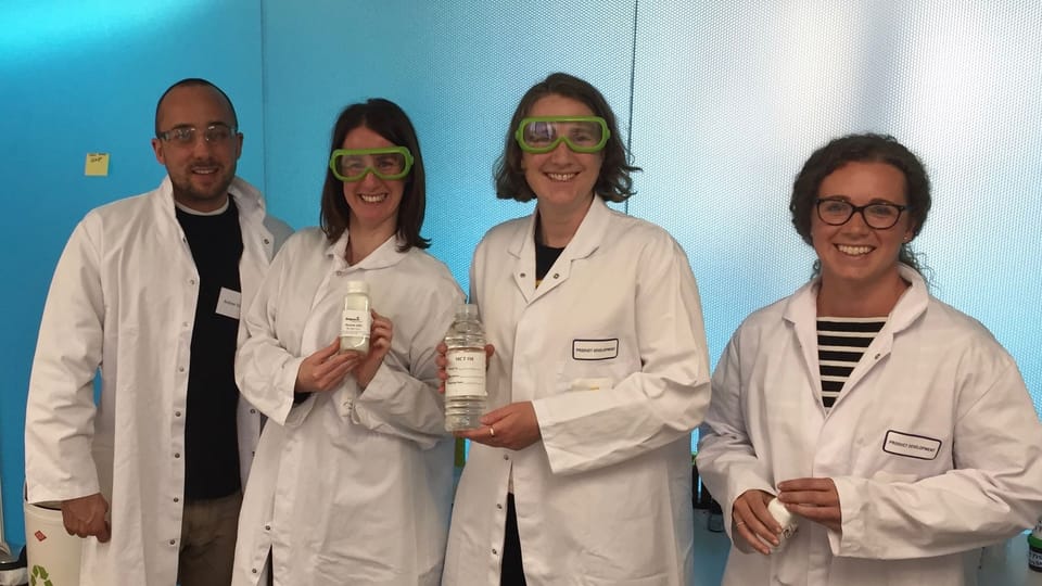 Four adults in lab coats and safety goggles standing together in a bright lab, smiling and holding bottles during a science activity.